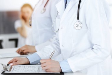 İş yerindeki bilinmeyen kadın doktorlar, arka planda hastaları var. Güneşli hastane resepsiyonunda beklerken tıbbi belgeleri dolduran kadın doktorlar, yakın plan. Sağlık hizmetleri