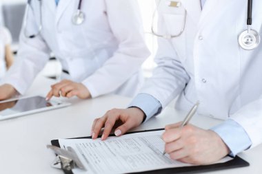 İş yerindeki bilinmeyen kadın doktorlar, arka planda hastaları var. Güneşli hastane resepsiyonunda beklerken tıbbi belgeleri dolduran kadın doktorlar, yakın plan. Sağlık hizmetleri