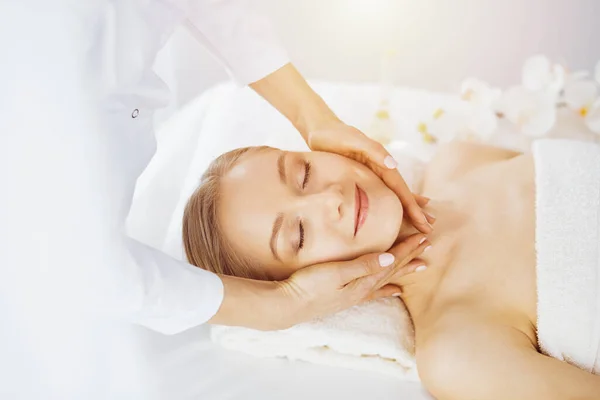 Beautiful caucasian woman enjoying facial massage with closed eyes in sunny spa salon. Relaxing treatment in medicine and Beauty concept — Stock Photo, Image