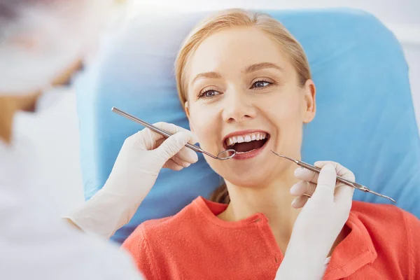 Uśmiechnięta biała kobieta jest badana przez dentystę w słonecznej klinice dentystycznej. Zdrowe zęby i medycyna, koncepcja stomatologii — Zdjęcie stockowe