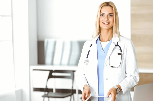 Mujer-médico en el trabajo en la clínica emocionada y feliz de su profesión. Mujer rubia médico está sonriendo mientras se utiliza la tableta. Concepto de medicina — Foto de Stock