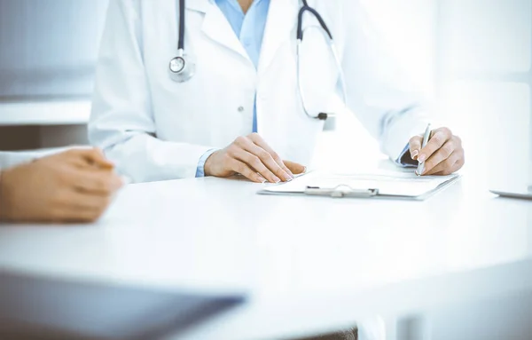 Mujer-médico desconocida y paciente sentada y hablando en el examen médico en la clínica, de cerca. El terapeuta que usa blusa azul está llenando el historial de medicamentos. Concepto de medicina — Foto de Stock