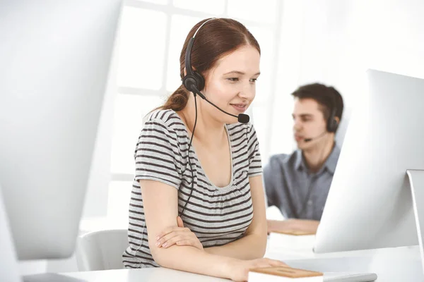 Casual dressed young woman using headset and computer while talking with customers online. Group of operators at work. Call center, business concept