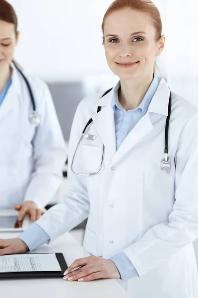Kvinna-läkare glad leende på jobbet med kollega och patient i bakgrunden. Porträtt av medelålders kvinnlig läkare fylla upp medicinska dokument eller recept när du står på sjukhus — Stockfoto