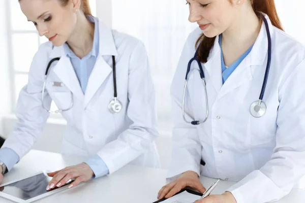 Two woman-doctors at work with patient at background. Female physicians filling up medical documents or prescription while standing in hospital reception desk. Data in medicine — Stock Photo, Image