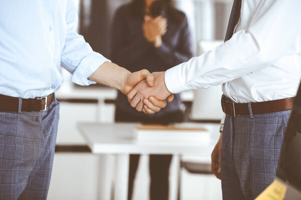 Two businessmen are shaking hands in office, close-up. Happy and excited business woman stands with raising hands at the background. Business people concept