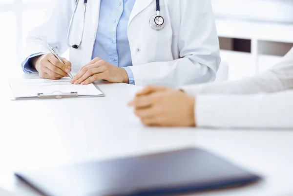 Mujer-médico desconocida y paciente sentada y hablando en el examen médico en la clínica, de cerca. El terapeuta que usa blusa azul está llenando el historial de medicamentos. Concepto de medicina — Foto de Stock