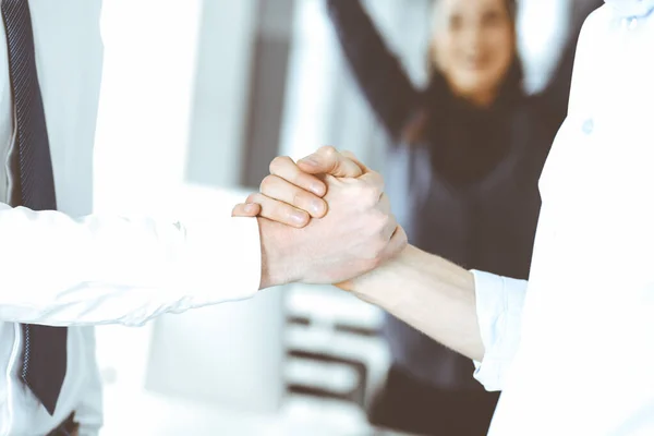 Dos hombres de negocios están estrechando las manos en la oficina, de cerca. Mujer de negocios feliz y emocionado se para con levantar las manos en el fondo. Concepto gente de negocios — Foto de Stock