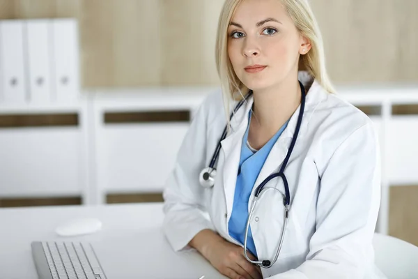 Портрет женщины-врача на работе, сидящей за столом в клинике. Блондинка веселый врач готов помочь пациентам — стоковое фото