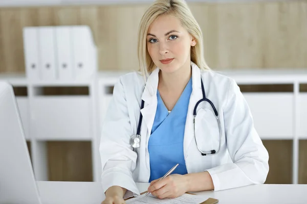 Nő-orvos portré munka közben ül az asztalnál a klinikán. Szőke, vidám orvos, aki kitölti az orvosi nyomtatványt vagy a receptet — Stock Fotó