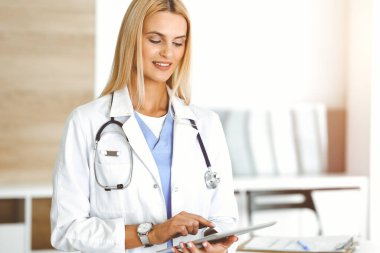 Kadın doktor, güneşli hastanede tablet bilgisayar kullanırken ilaç geçmişi ve sınav sonuçlarını kontrol ediyor. Tıp ve sağlık hizmetleri kavramı