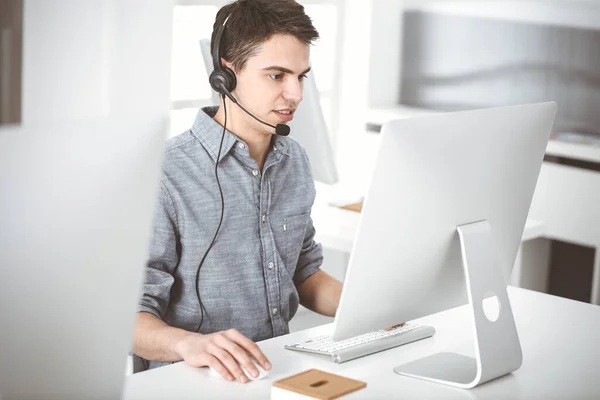 Casual dressed young man using headset and computer while talking with customers online. Call center, business concept