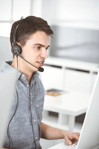 Hombre joven vestido casual usando auriculares y computadora mientras habla con los clientes en línea. Call center, concepto de negocio —  Fotos de Stock