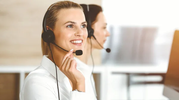 Mujer rubia representante de servicio al cliente está consultando a los clientes en línea utilizando auriculares en la oficina soleada. Concepto de Call center — Foto de Stock