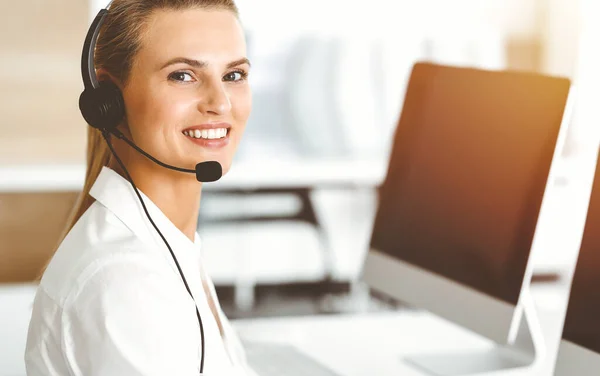 Mujer rubia representante de servicio al cliente está consultando a los clientes en línea utilizando auriculares en la oficina soleada. Concepto de Call center — Foto de Stock