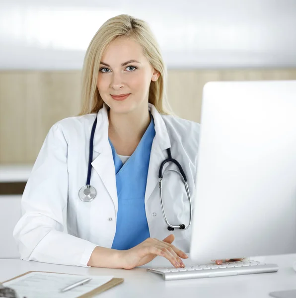 Mulher-médico no trabalho enquanto sentado na mesa no hospital ou na clínica. Médico alegre loira usando computador desktop — Fotografia de Stock