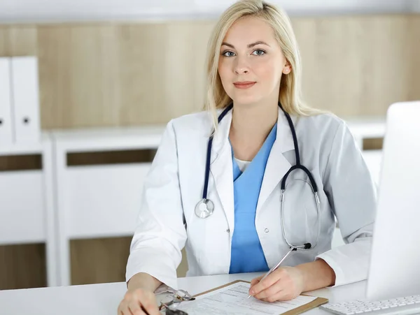 Retrato de mulher-médico no trabalho enquanto sentado na mesa na clínica. Médico alegre loiro preenchendo formulário médico ou prescrição — Fotografia de Stock