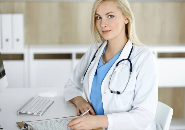 Retrato de mujer-médico en el trabajo mientras está sentado en el escritorio en la clínica. Rubia médico alegre llenando formulario médico o prescripción — Foto de Stock