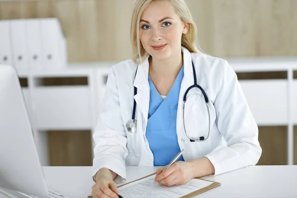 Portrato di donna-medico al lavoro seduto alla scrivania in clinica. Bionda allegro medico riempiendo modulo medico o prescrizione — Foto Stock
