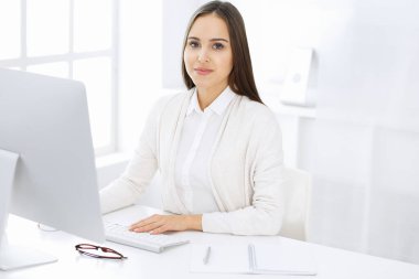 Genç, neşeli bir kadın bilgisayarla masada oturuyor ve beyaz renkli ofisteki kameraya bakıyor. Öğrenci kız ya da iş kadınına benziyor. Vesikalık