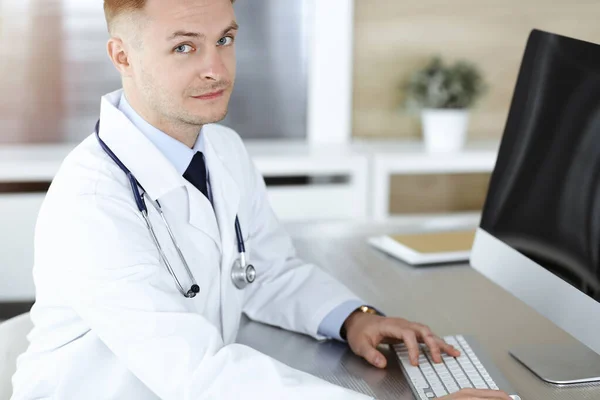 Doctor hombre sentado detrás de una computadora en el lugar de trabajo en la clínica soleada. — Foto de Stock