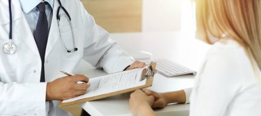 Doktor ve hasta, güneşli klinikteki masada otururken tıbbi muayene sonuçlarını tartışıyorlar. Yakın plan. Erkek doktor pano kullanıyor.