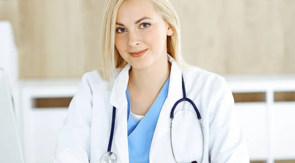 Retrato de mulher-médico no trabalho enquanto sentado na mesa na clínica. Médico alegre loiro preenchendo formulário médico ou prescrição — Fotografia de Stock