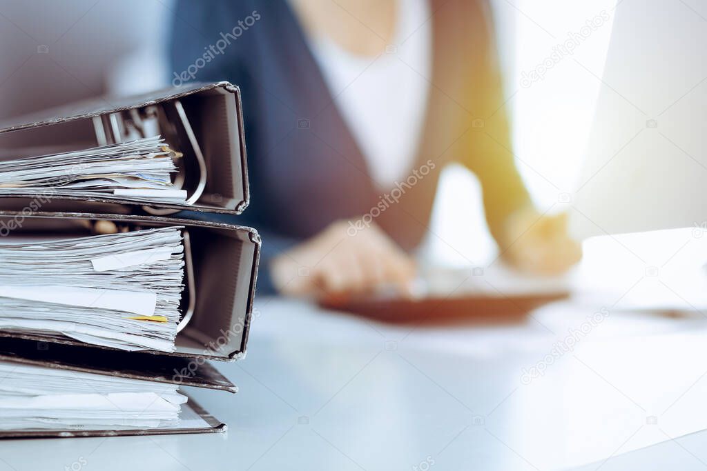 Binders with papers are waiting to be processed by business woman or bookkeeper working at the desk in office back in blur. Audit and tax concept