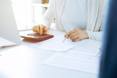 Muhasebeci mali durumu kontrol ediyor ya da vergi formları için hesap makinesi geliri ile sayıyor, eller yakın çekim. İş kadını ofisteki masada iş arkadaşıyla oturuyor ve çalışıyor. Denetim kavramı