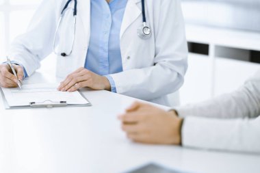 Bilinmeyen kadın-doktor ve kadın hasta klinikteki tıbbi muayenede oturuyor ve konuşuyor, yakın plan. Mavi bluz giyen terapist ilaç geçmişini dolduruyor. Tıp konsepti