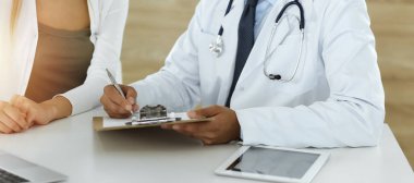 Doktor ve hasta, güneşli klinikteki masada otururken tıbbi muayene sonuçlarını tartışıyorlar. Yakın plan. Erkek doktor pano kullanıyor.
