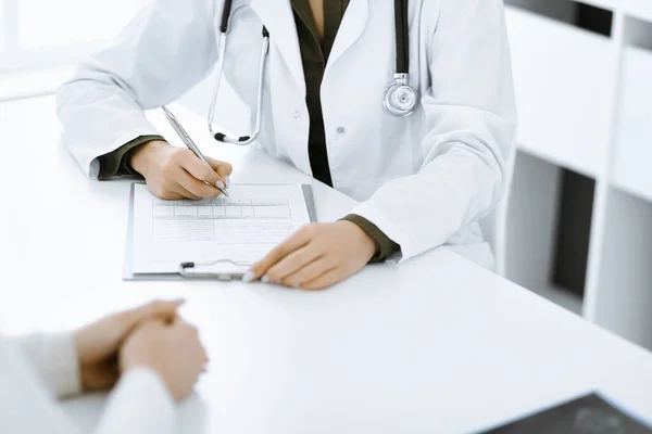 Onbekende vrouw-dokter en vrouwelijke patiënt zitten en praten bij medisch onderzoek in de kliniek, close-up. Therapeut met groene blouse vult de geschiedenis van medicatie. Geneesmiddelenconcept — Stockfoto