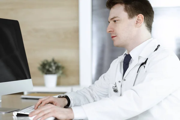 Doctor hombre sentado detrás de una computadora en el lugar de trabajo en la clínica u oficina del hospital. Fotografía médica y concepto de medicina — Foto de Stock