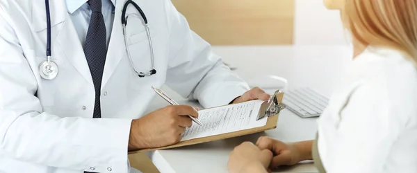 Doctor and patient discussing medical exam results while sitting at the desk in sunny clinic, close-up. Male physician using clipboard.
