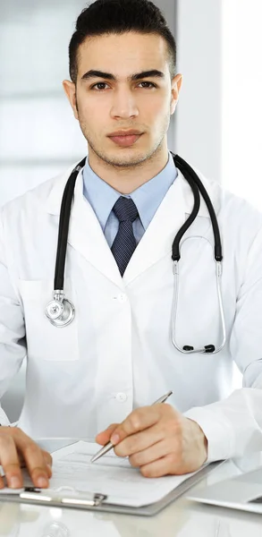 Arabischer Arzt benutzt Laptop-Computer, während er am gläsernen Schreibtisch in der Klinik das Formular mit der Medikamentengeschichte ausfüllt. Medizinkonzept — Stockfoto