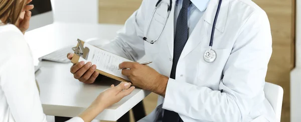 Dokter en patiënt bespreken medische onderzoeksresultaten terwijl ze aan de balie zitten in kliniek, close-up. Mannelijke arts met behulp van klembord voor het vullen van medicatie geschiedenis record van jonge vrouw — Stockfoto