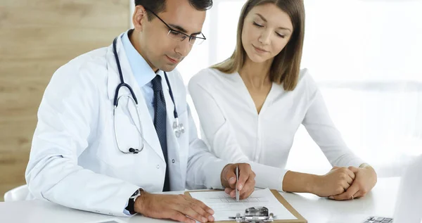 Doctor and patient discussing medical exam results while sitting at the desk in clinic. Male physician using clipboard for filling up medication history record of young woman. Data in medicine
