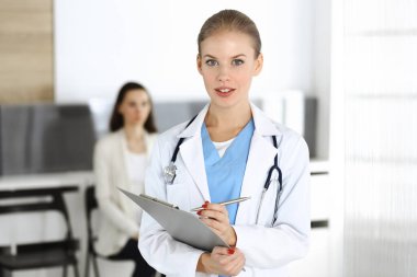 Kadın - doktor hastanın arka planında klinikte tıbbi geçmiş kayıtlarını doldurmak için pano kullanıyor. Tıp konsepti