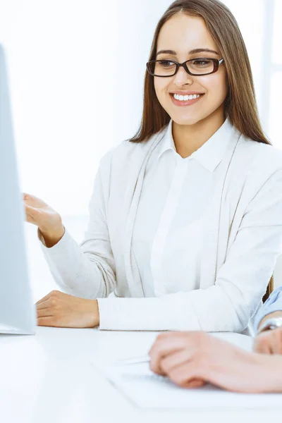 Mujer joven sentada en el escritorio con computadora en la oficina de color blanco. Parece estudiante o dama de negocios comunicándose con el hombre vestido casual —  Fotos de Stock