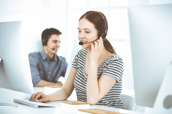 Casual dressed young woman using headset and computer while talking with customers online. Group of operators at work. Call center, business concept