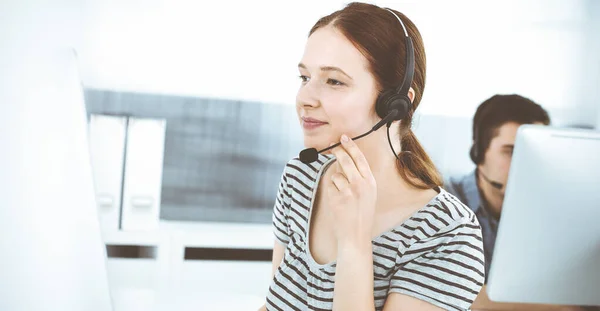 在网上与客户交谈时，用耳机和电脑随意穿衣的年轻女性。一组操作员在工作。呼叫中心，商业概念 — 图库照片
