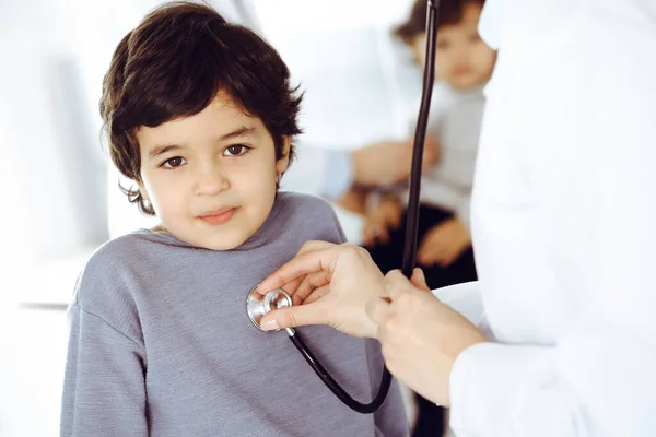 Doctor-woman examining a child patient by stethoscope. Cute arab boy and his brother at physician appointment. Medicine concept — Stock Photo, Image