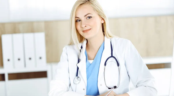 Mujer-médico en el trabajo mientras está sentado en el escritorio en el hospital o la clínica. Rubia alegre médico llenar la historia de la medicación formulario de registro — Foto de Stock