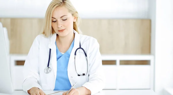 Mujer-médico en el trabajo mientras está sentado en el escritorio en el hospital o la clínica. Rubia alegre médico llenar la historia de la medicación formulario de registro — Foto de Stock