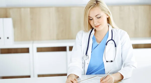 Женщина-врач на работе, сидя за столом в больнице или клинике. Блондинка, веселый врач, заполняет анкету с лекарствами — стоковое фото