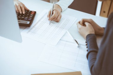 Muhasebeci mali durumu kontrol ediyor ya da vergi formları için hesap makinesi geliri ile sayıyor, eller yakın çekim. İş kadını ofisteki masada iş arkadaşıyla oturuyor ve çalışıyor. Denetim kavramı