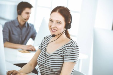 İnternette müşterilerle konuşurken kulaklık ve bilgisayar kullanan sıradan giyinmiş genç bir kadın. Bir grup operatör iş başında. Çağrı merkezi, iş konsepti