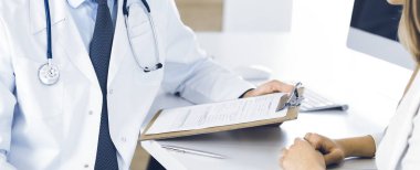 Doktor ve hasta klinikteki masada otururken tıbbi muayene sonuçlarını tartışıyorlar, yakın plan. Genç bir kadının ilaç geçmişini doldurmak için pano kullanan bir erkek doktor.