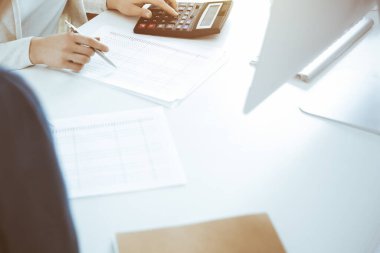 Muhasebeci mali durumu kontrol ediyor ya da vergi formları için hesap makinesi geliri ile sayıyor, eller yakın çekim. İş kadını ofisteki masada iş arkadaşıyla oturuyor ve çalışıyor. Denetim kavramı