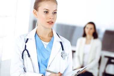 Kadın - doktor hastanın arka planında klinikte tıbbi geçmiş kayıtlarını doldurmak için pano kullanıyor. Tıp konsepti